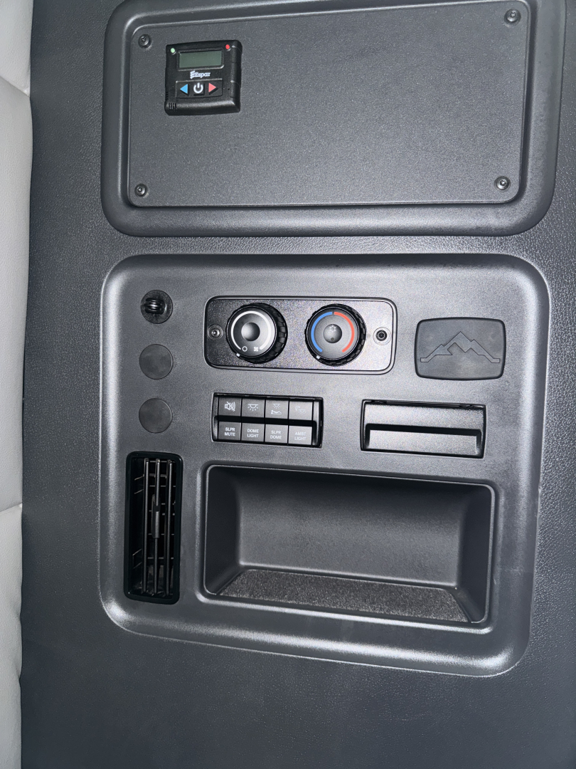 Cab interior panel of a Freightliner Cascadia showing climate control dials, a storage pocket, and an auxiliary panel with buttons; upper compartment panel visible.