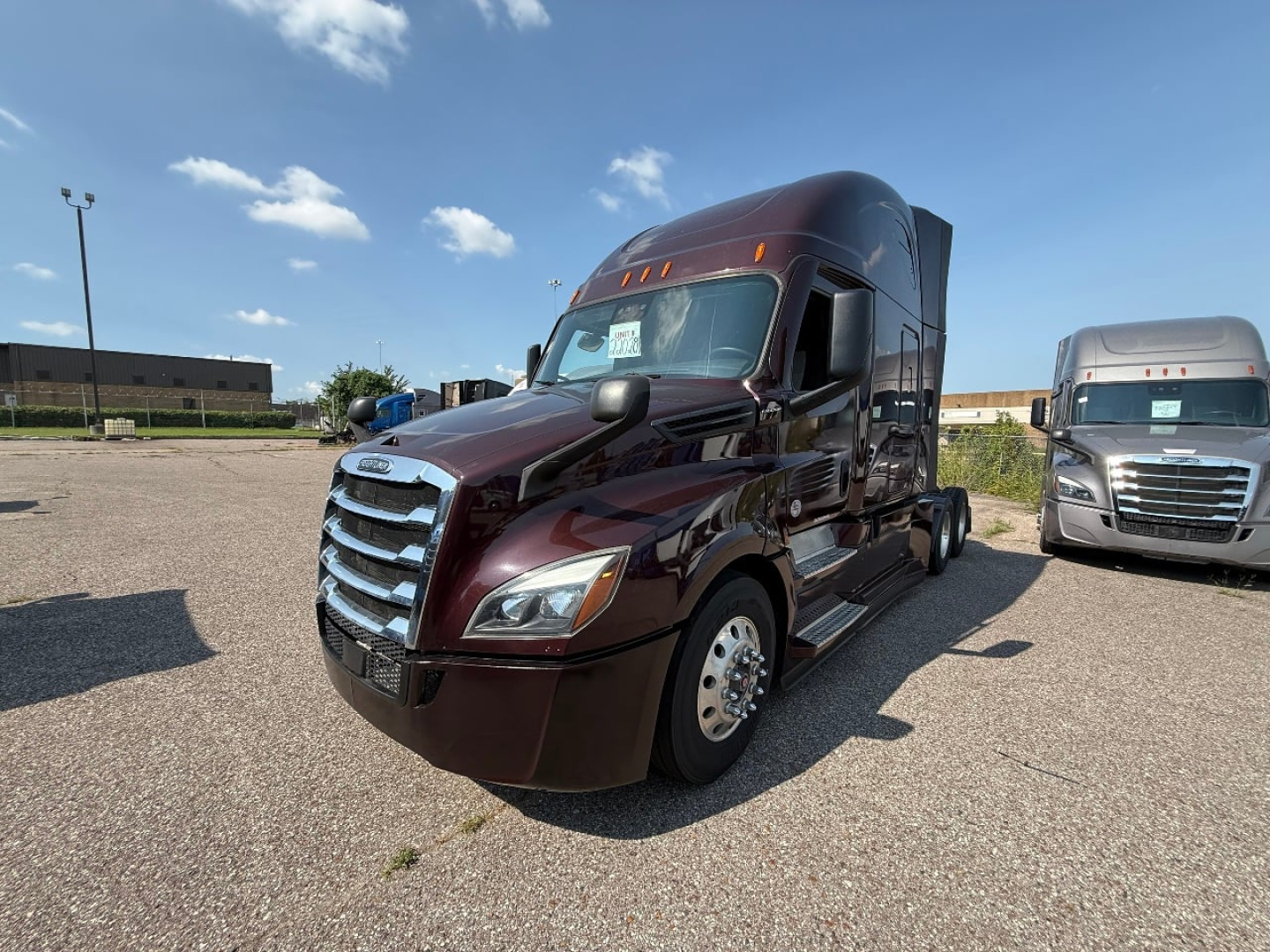 Conventional sleeper truck in a parking lot showing a maroon 2022 Freightliner Cascadia 126 tractor with chrome grille and side steps; adjacent light-colored truck in background.