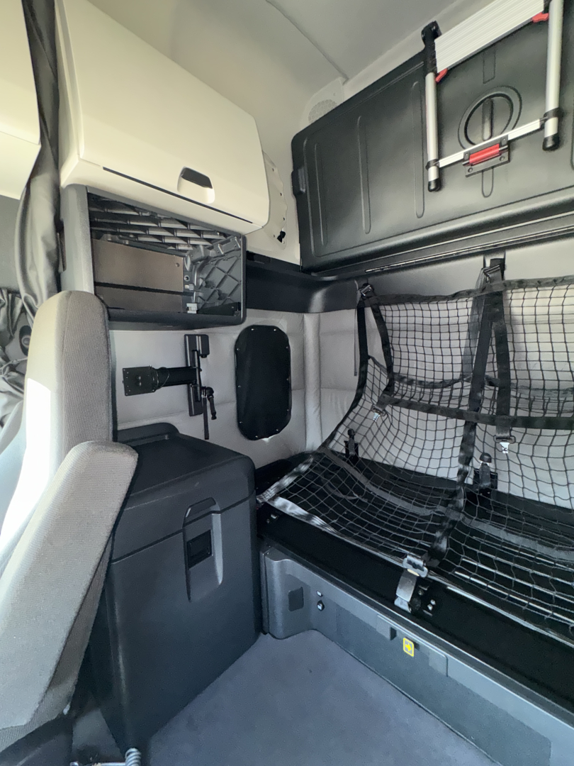 Truck cab interior in a Freightliner Cascadia sleeper. Visible storage compartments, netted bunk area with safety net, cabinet, and chair. Includes mesh guard and overhead equipment.