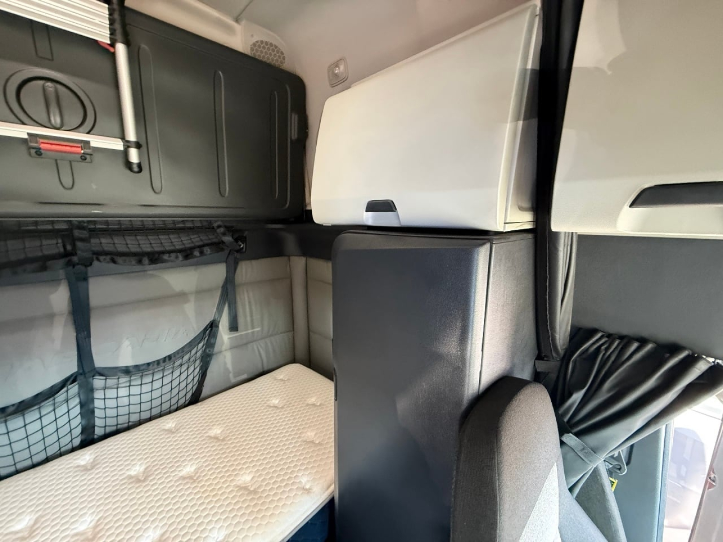 Cab interior of a Freightliner Cascadia sleeper showing bunk bed area, storage nets, overhead cabinet, and window curtain.