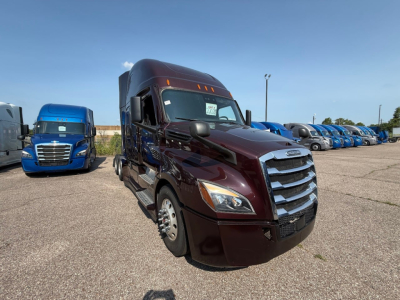 Truck front view in a parking lot showing Freightliner Cascadia cab with maroon color, chrome grille, headlights, and side mirrors; other blue trucks in background.