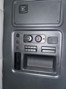Cab interior panel of a Freightliner Cascadia showing climate control dials, a storage pocket, and an auxiliary panel with buttons; upper compartment panel visible.