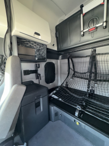 Truck cab interior in a Freightliner Cascadia sleeper. Visible storage compartments, netted bunk area with safety net, cabinet, and chair. Includes mesh guard and overhead equipment.