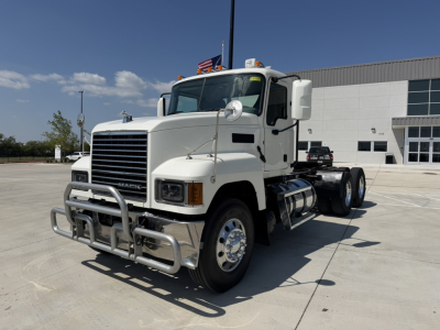 White Mack PI64T conventional daycab truck in front of a dealership building showing front grille with bumper guard, large mirrors, dual rear wheels, and fuel tanks; no legible small text visible.