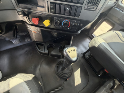 Truck cab interior with manual gear shifter and dashboard controls. Visible text on gear knob reads MACK with gear numbers. Dashboard has climate controls, red, yellow, and black knobs, and various buttons.