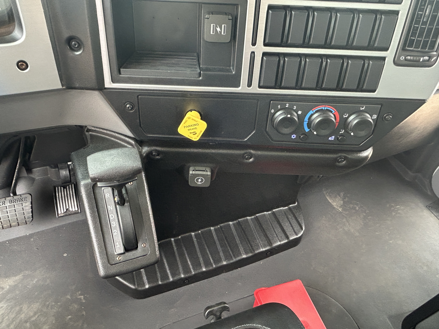 Mack MD6 cab interior showing dash controls, climate knobs, and a yellow hood release handle with truck floor mat and pedals.