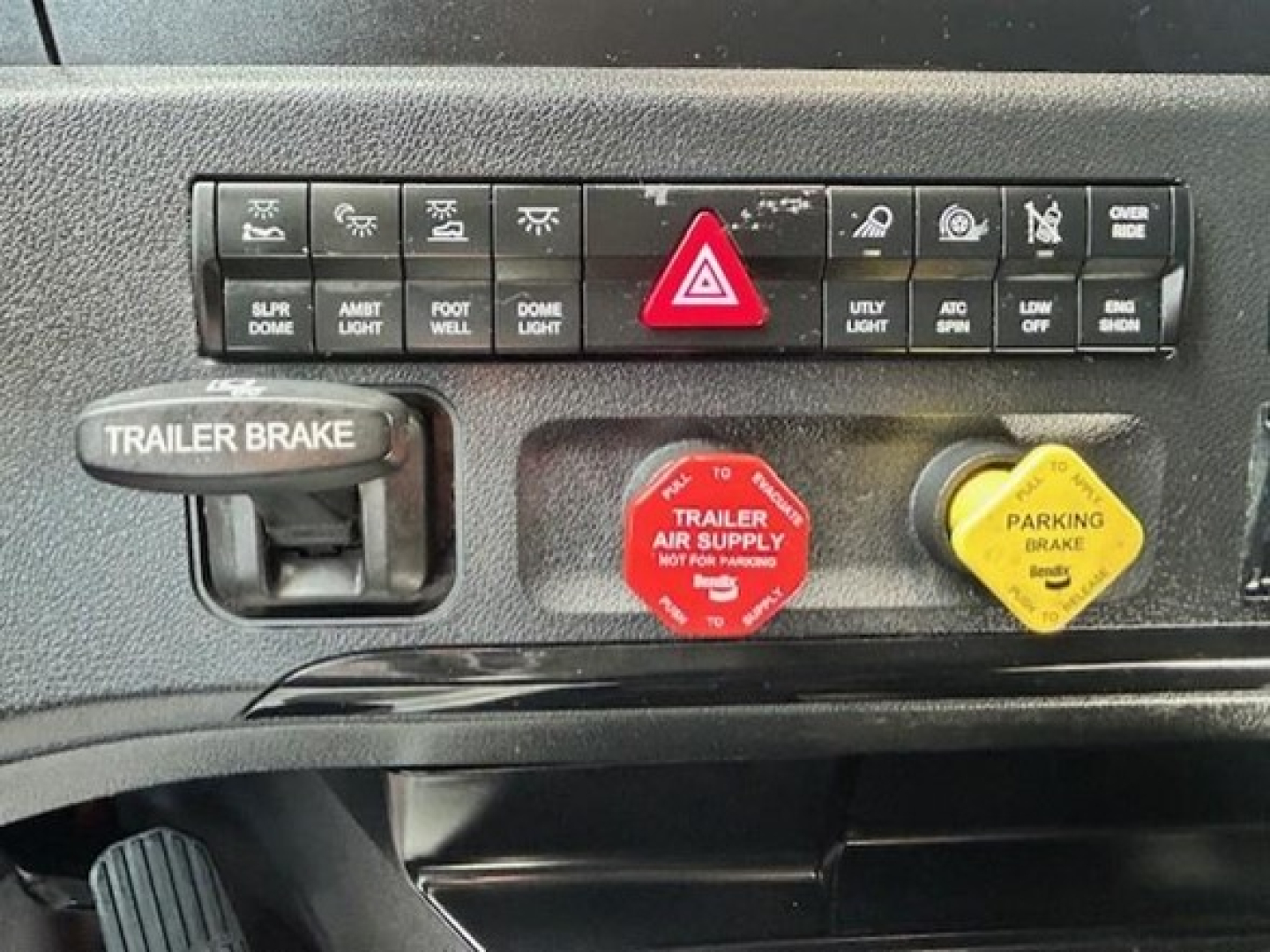 Dashboard controls and emergency switches in a Freightliner Cascadia cabin: red hazard button, red trailer air supply WARNING, yellow parking brake, trailer brake lever, row of small indicator buttons.