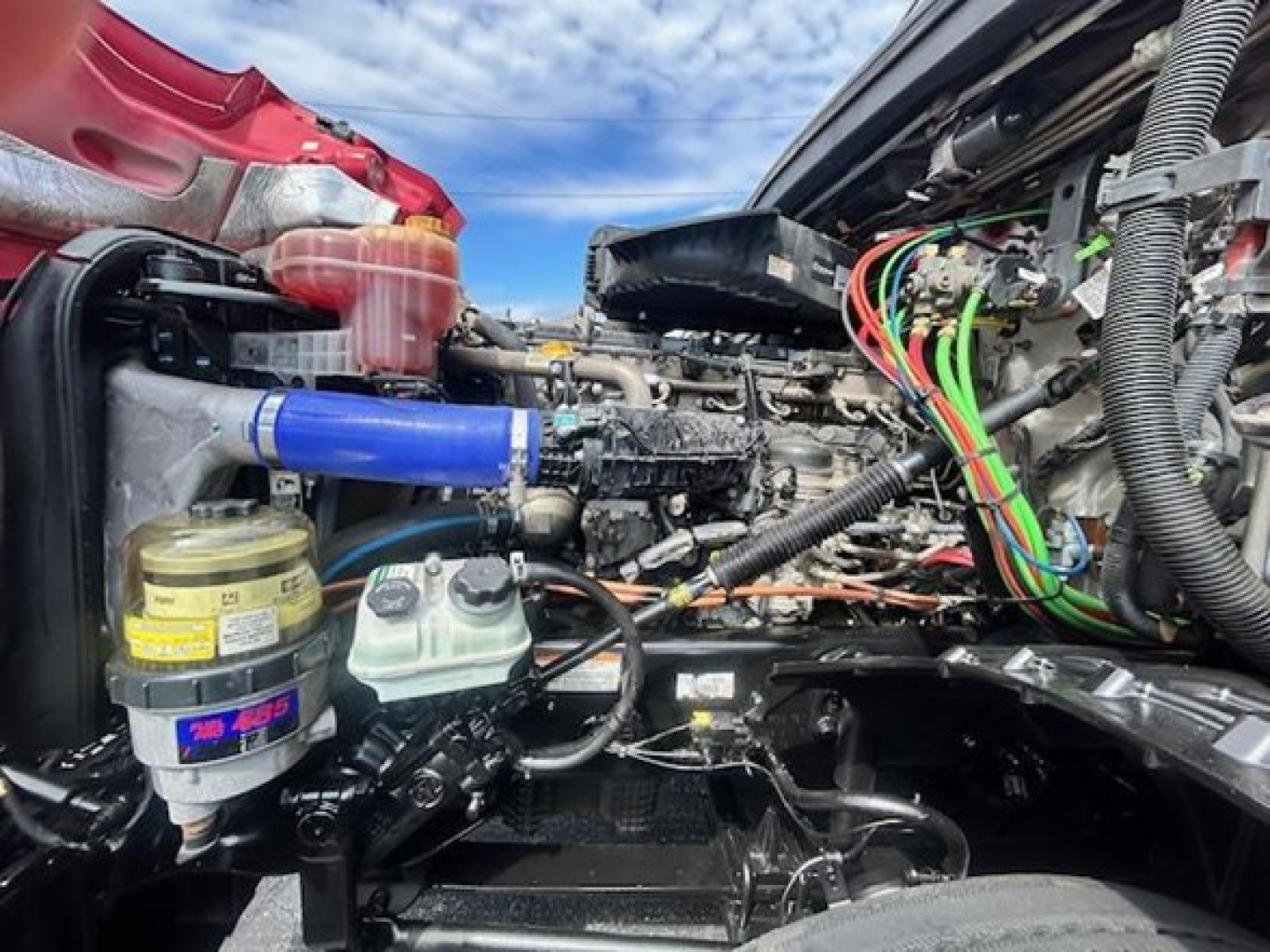 Truck engine bay with various colored hoses and wiring harnesses visible; brake fluid reservoir, coolant reservoir, and diesel engine components seen in a Freightliner Cascadia truck engine bay.