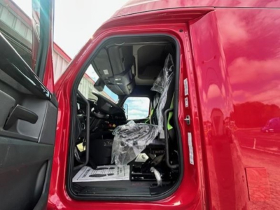 Cabin of a red Freightliner Cascadia 126 truck with open driver door, plastic-wrapped seat, and dashboard visible.