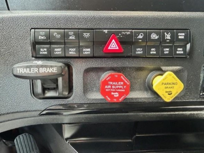 Dashboard controls and emergency switches in a Freightliner Cascadia cabin: red hazard button, red trailer air supply WARNING, yellow parking brake, trailer brake lever, row of small indicator buttons.