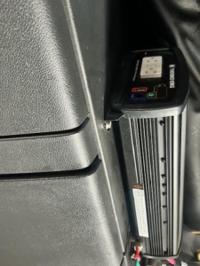 Power inverter mounted in cab with visible red and blue DC connectors and label, mounted beside dashboard panel in Freightliner Cascadia interior.