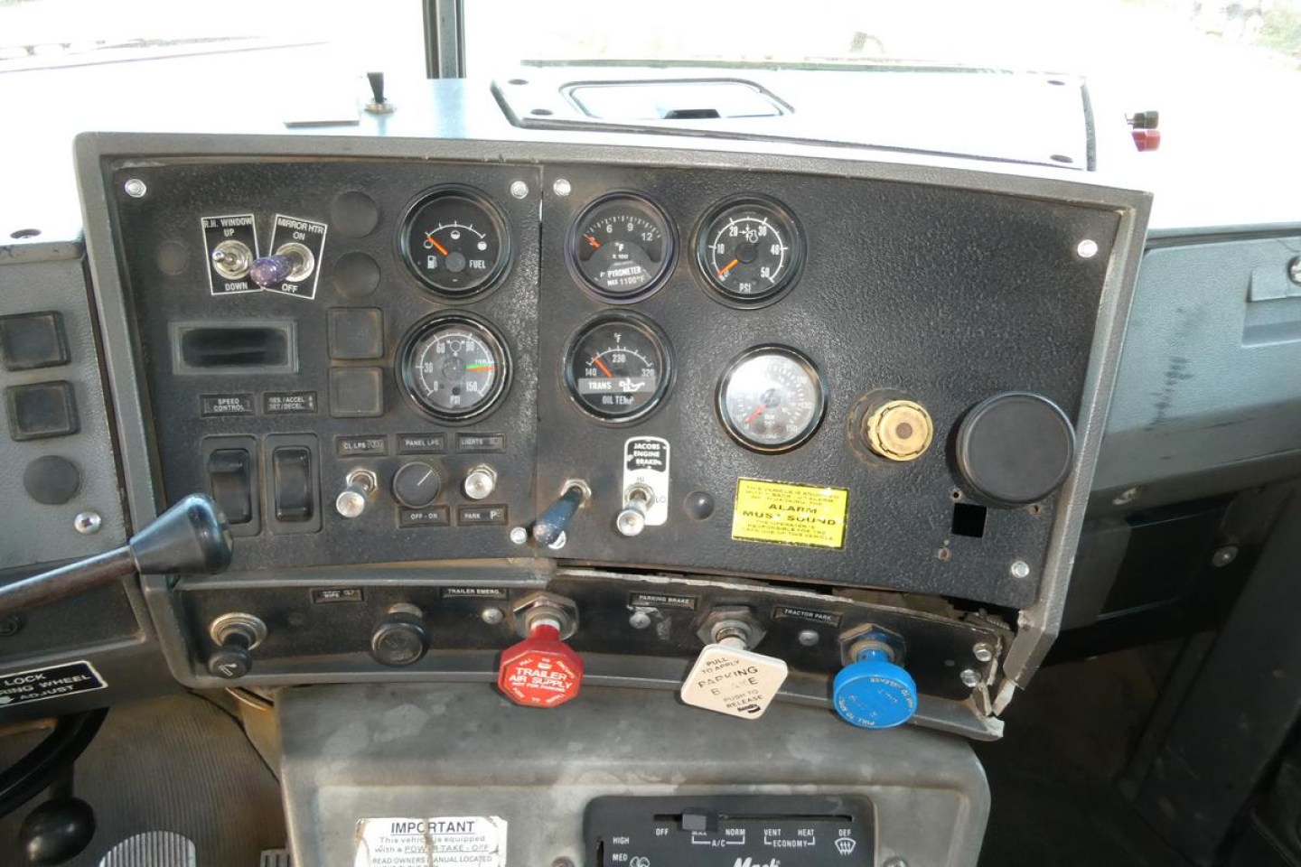 Truck dashboard with multiple gauges and control switches. Visible text includes labels near knobs and a yellow warning plate. Red, white, and blue knobs at bottom with text.