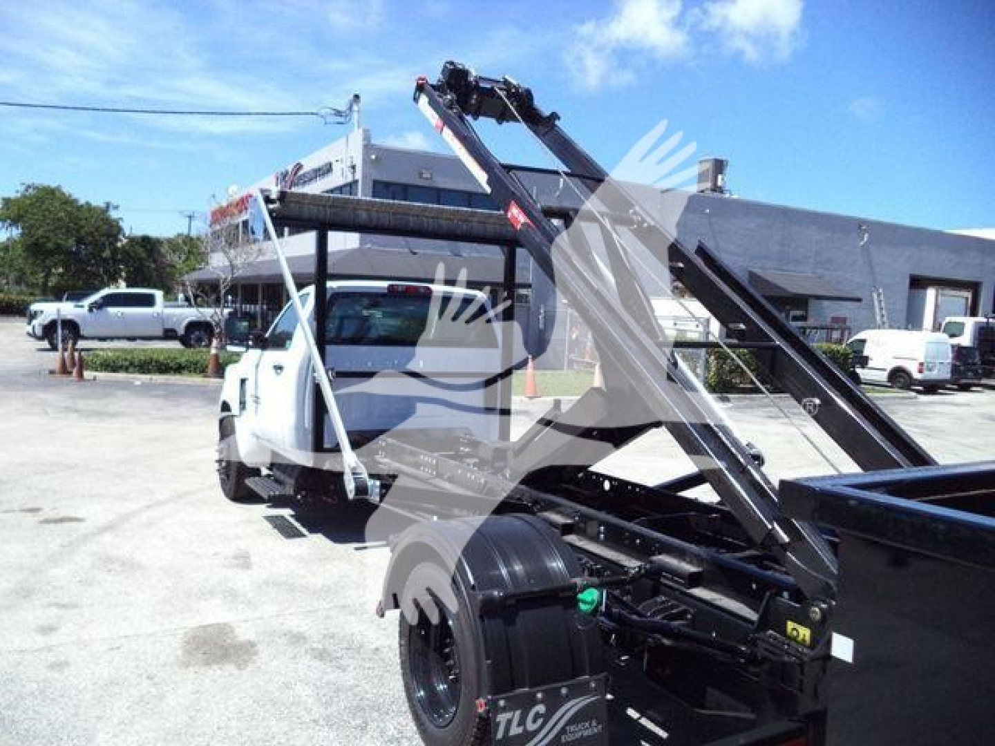 Truck chassis and hydraulic arm lift attached to a white Chevrolet Silverado 6500HD roll-off truck in a dealership lot.