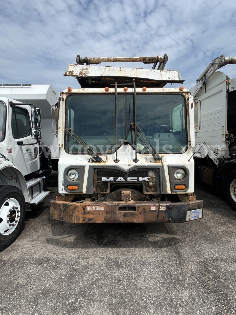 Front view of a 2012 Mack MRU613 trash truck showing worn front bumper and rusted boom on top; Mack grille with logo visible.