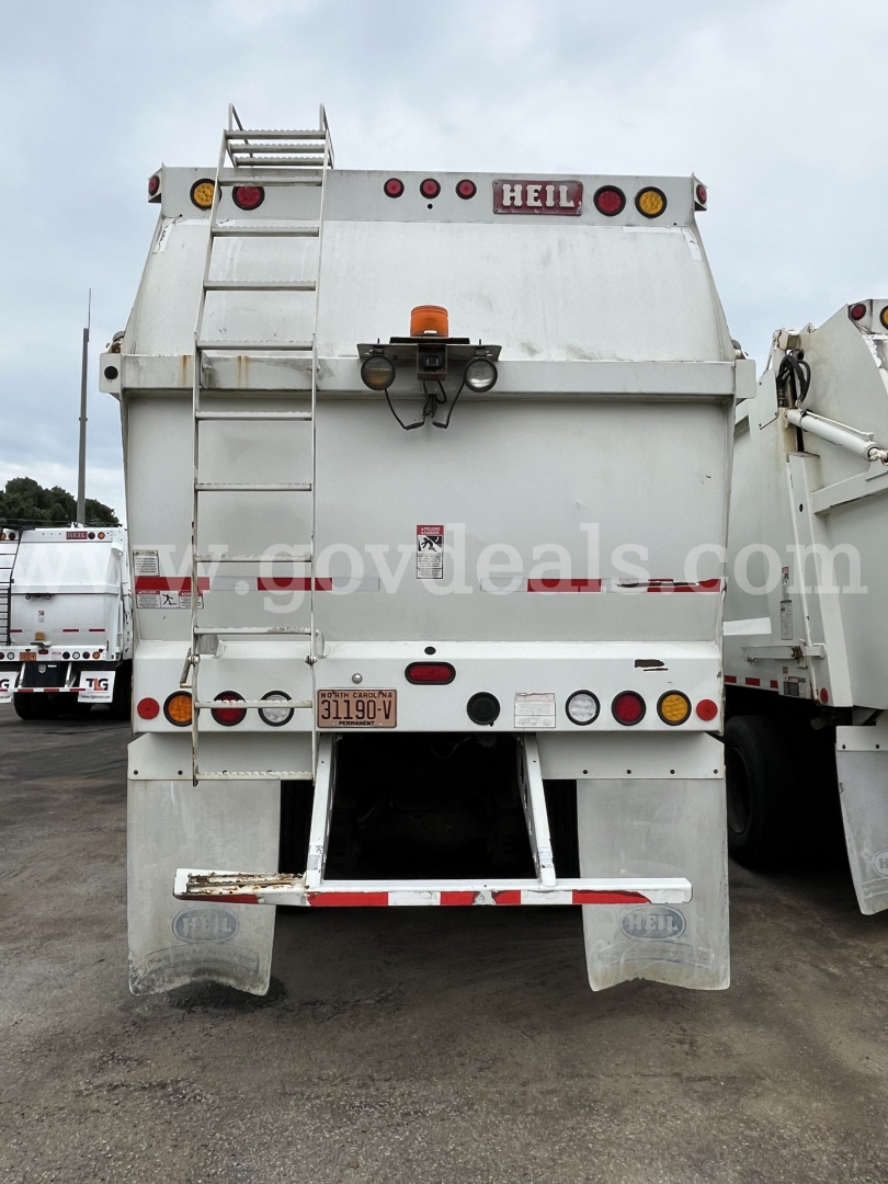 Rear view of a white 2012 Mack MRU613 refuse truck with HEIL branding, ladder on left, multiple lights, and a double tailgate.