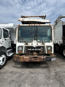 Front view of a 2012 Mack MRU613 trash truck showing worn front bumper and rusted boom on top; Mack grille with logo visible.