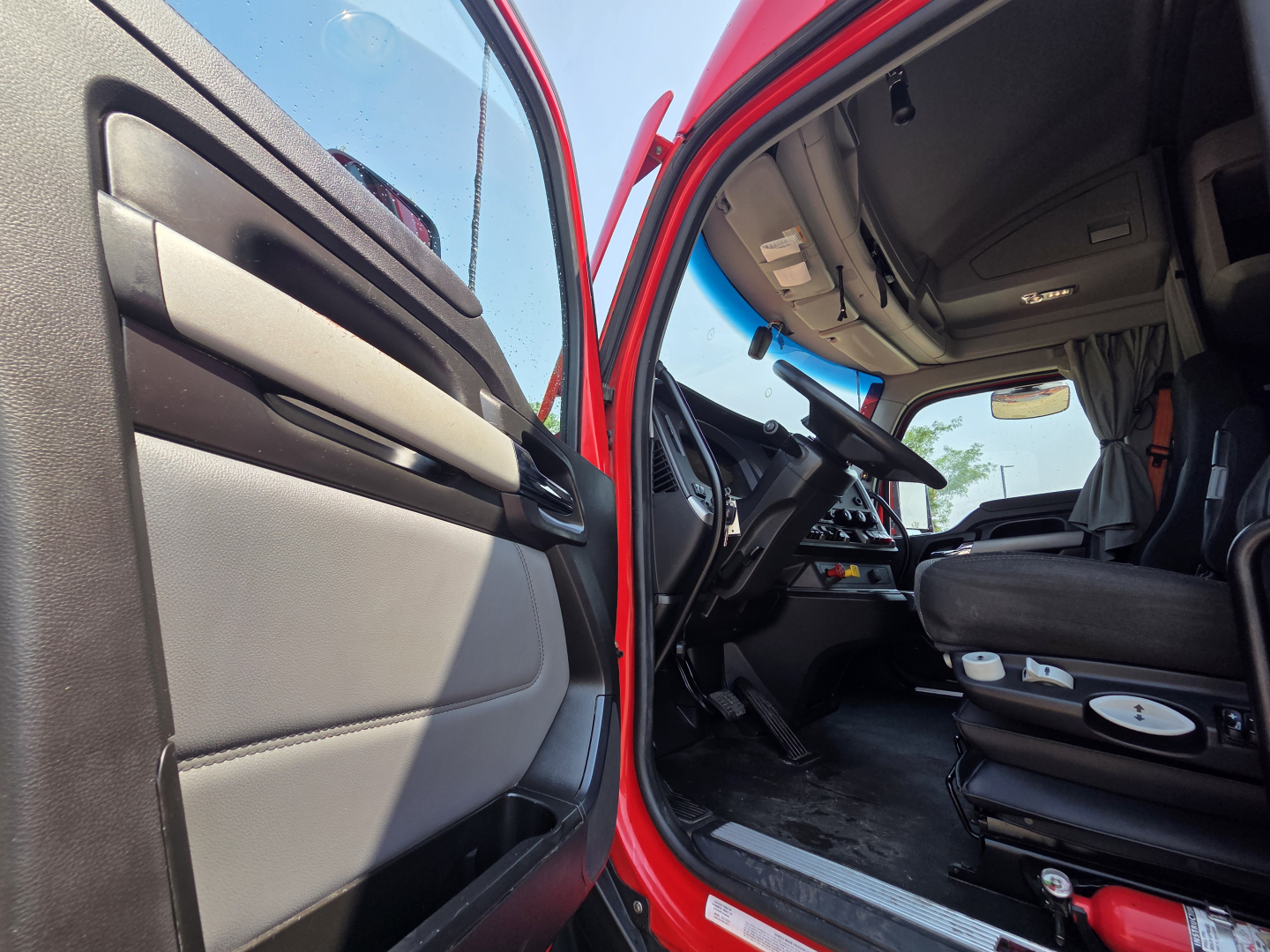 Truck cab interior of a red 2023 Kenworth T680 showing the driver seat, steering wheel, and open door, VIN plate area not legible.