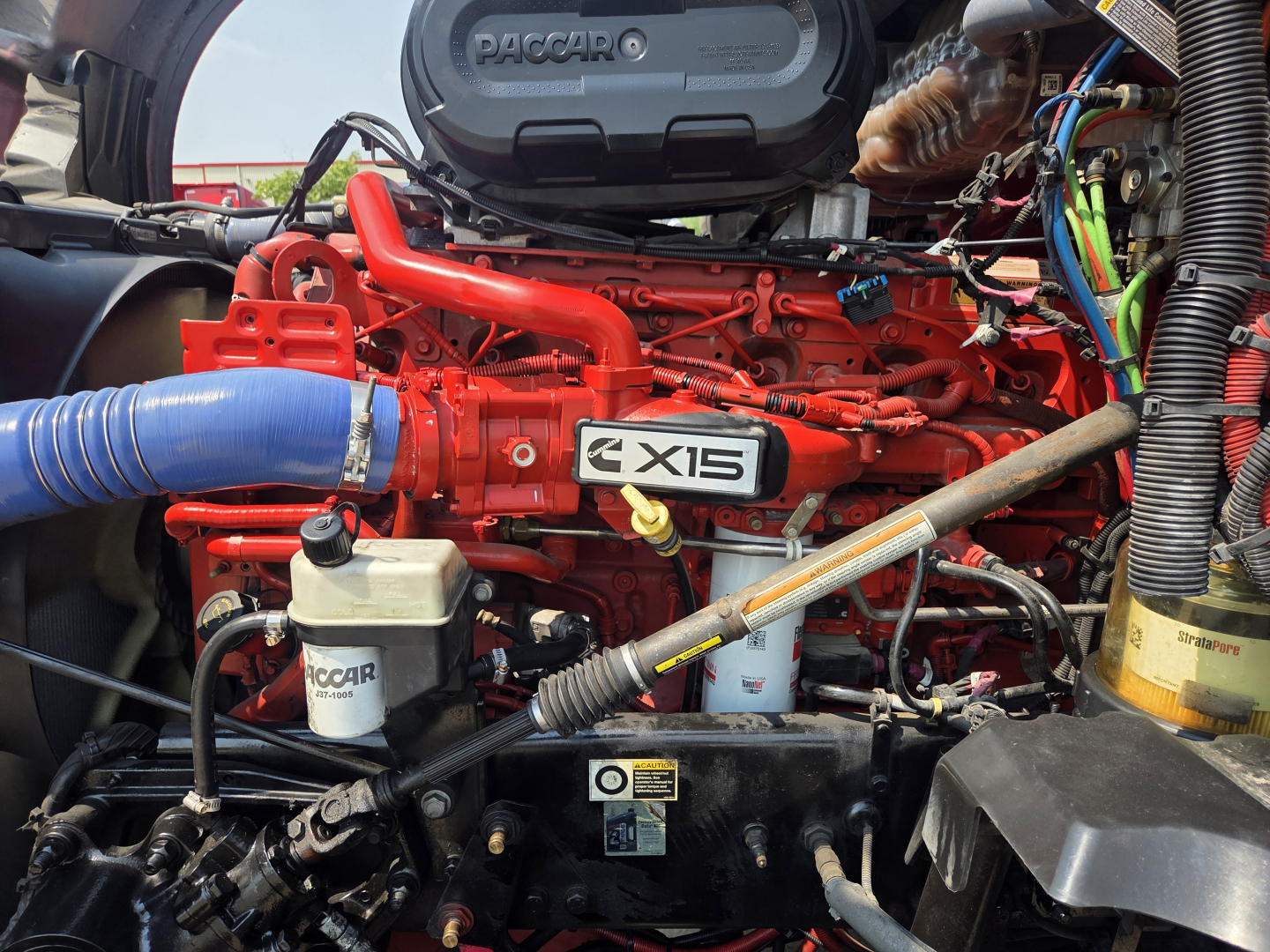 Kenworth T680 engine bay showing PacCar i6 15? engine with red block and blue intercooler hose, various wiring and fluid reservoirs.
