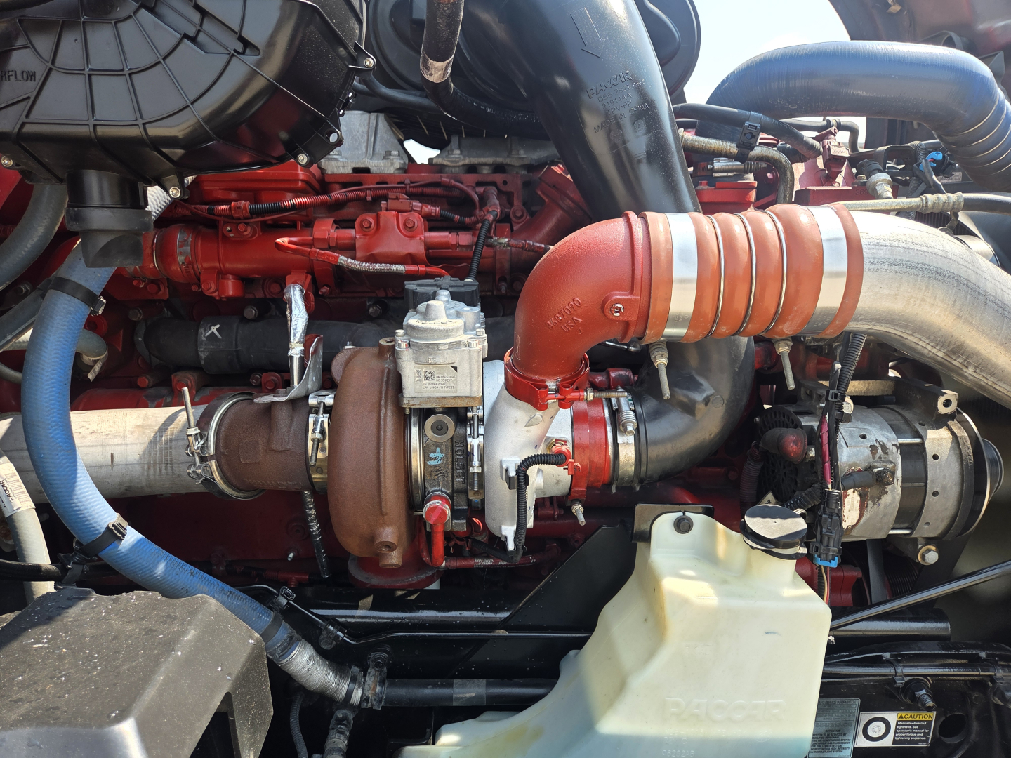 Kenworth T680 engine bay showing turbocharger lines and red engine block with orange intercooler hose and coolant reservoir.