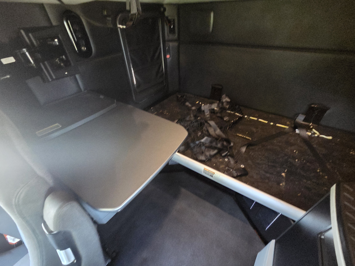Interior of a 2023 Kenworth T680 truck cab with a fold-out table and a bed area showing a dark surface and some wiring. No legible labels visible.