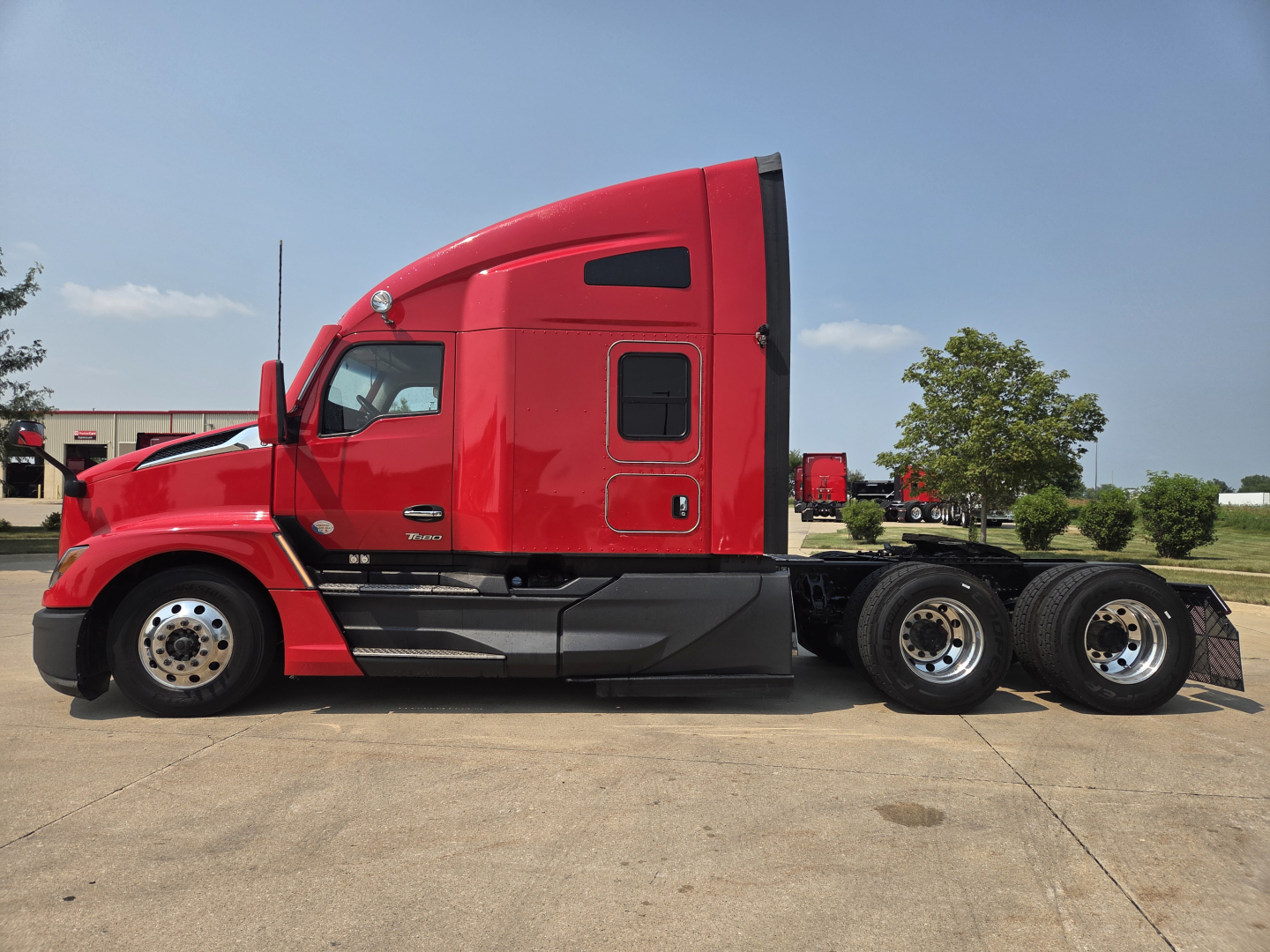 Kenworth T680 tractor cab side view showing red exterior, door with window, fuel/equipment compartments, and triple rear axles with chrome wheels.