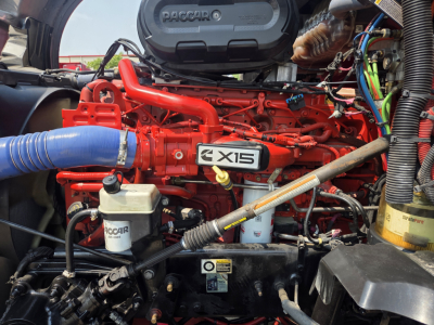 Kenworth T680 engine bay showing PacCar i6 15? engine with red block and blue intercooler hose, various wiring and fluid reservoirs.