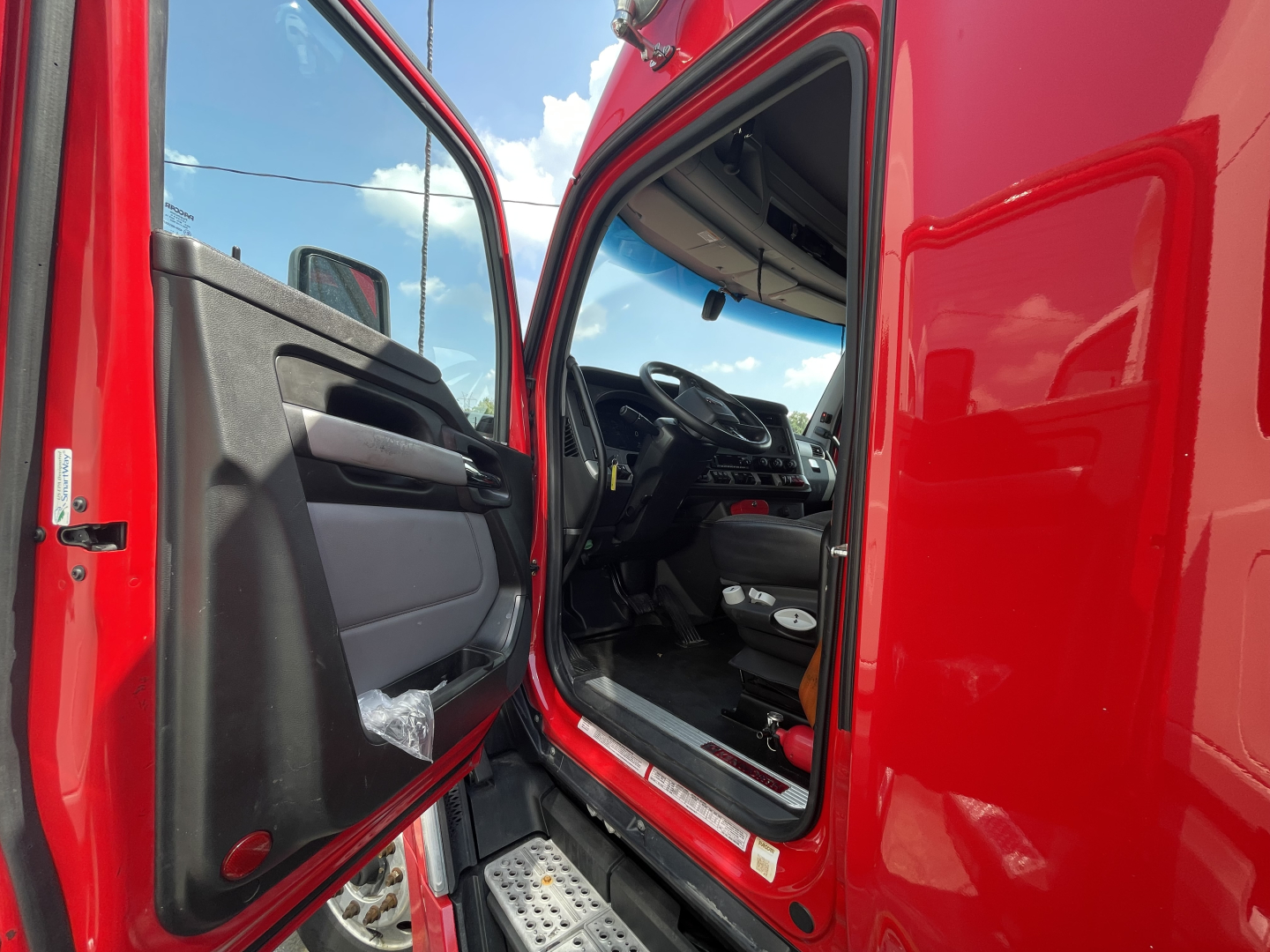Truck cab interior view with open door showing steering wheel and dashboard of a red 2022 Kenworth T680, front seats visible.