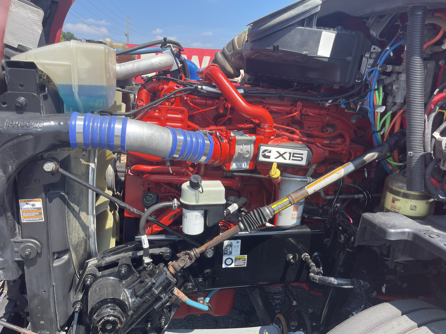 Kenworth T680 engine bay showing CE15 engine with red intake manifold, blue silicone turbo boot, fuel filters, hoses, and wiring harnesses.