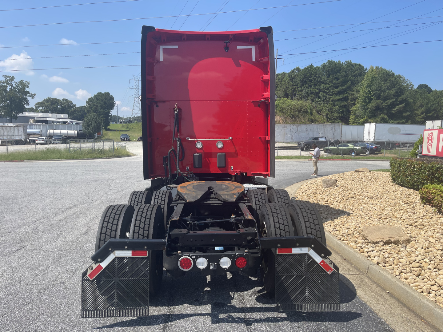 Kenworth T680 chassis rear view showing dual rear axles and red sleeper cab doors open, with mud flaps and hydraulic lines; rear lights and connectors visible.
