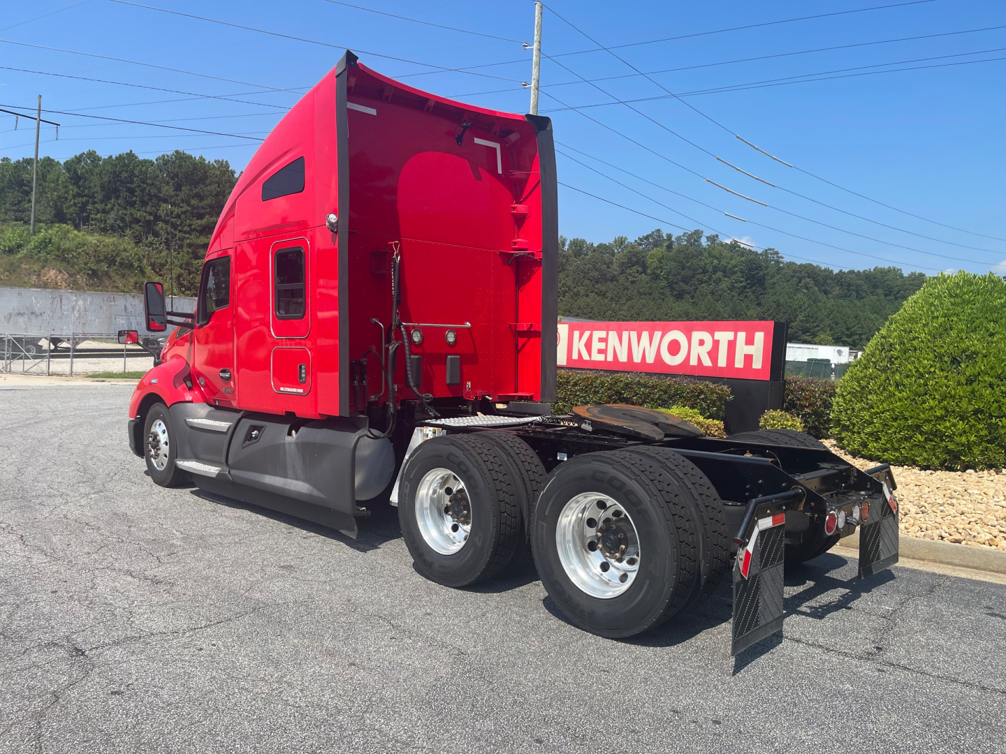 Kenworth T680 sleeper cab tractor (rear view) showing triple rear axles, red cab, black chassis, Kenworth dealership sign in background