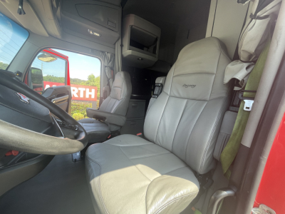 Cab interior of a Kenworth truck showing driver seat with 'Dynaflex' logo? and overhead storage; steering wheel and dashboard visible.