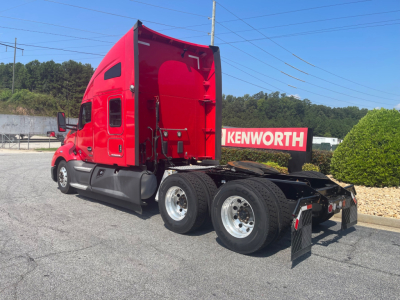 Kenworth T680 sleeper cab tractor (rear view) showing triple rear axles, red cab, black chassis, Kenworth dealership sign in background
