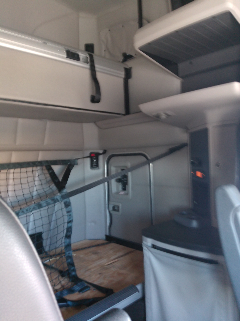 Interior of a Peterbilt truck cab showing sleeper area, overhead storage compartments, and a partition with a safety net; cockpit area visible on left.