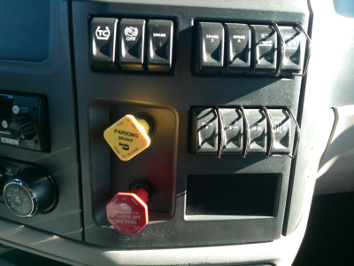 Truck cab dashboard controls with yellow parking brake knob and red stop/safety knob visible; multiple rocker switches and gauges.