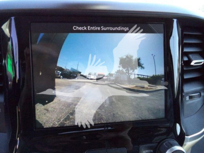 In-vehicle touchscreen display showing rear camera view with 'Check Entire Surroundings' text and reflection of the parking lot.