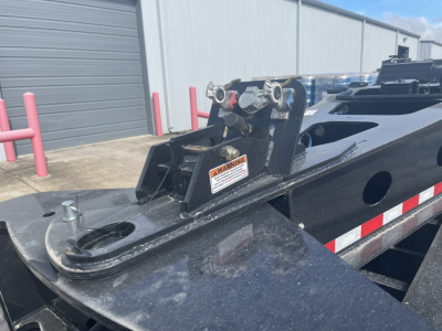 Trailer hitch and mounting hardware on Load King SB141/2-8'-6" trailer, black steel, warning sticker, reflected red/white stripe, industrial yard background.