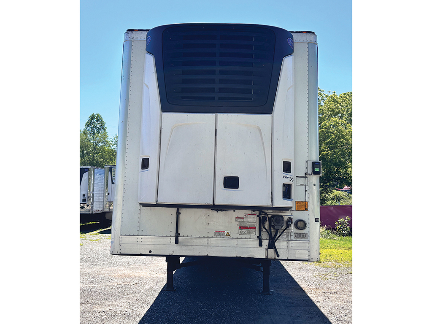 Rear view of a utility reefer trailer with a black top hatch and white doors, showing safety decals and cables near the bumper.