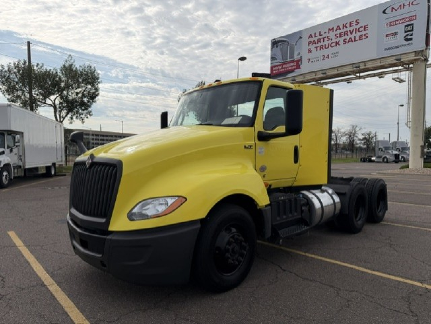 Yellow 2023 International LT625 cab and chassis truck in a parking lot with a signboard in background. Visible text includes VIN 3HSDZAPR0PN789552 on listing, and a billboard reading ALL MAKES PARTS, SERVICE & TRUCK SALES, with a logo. No e