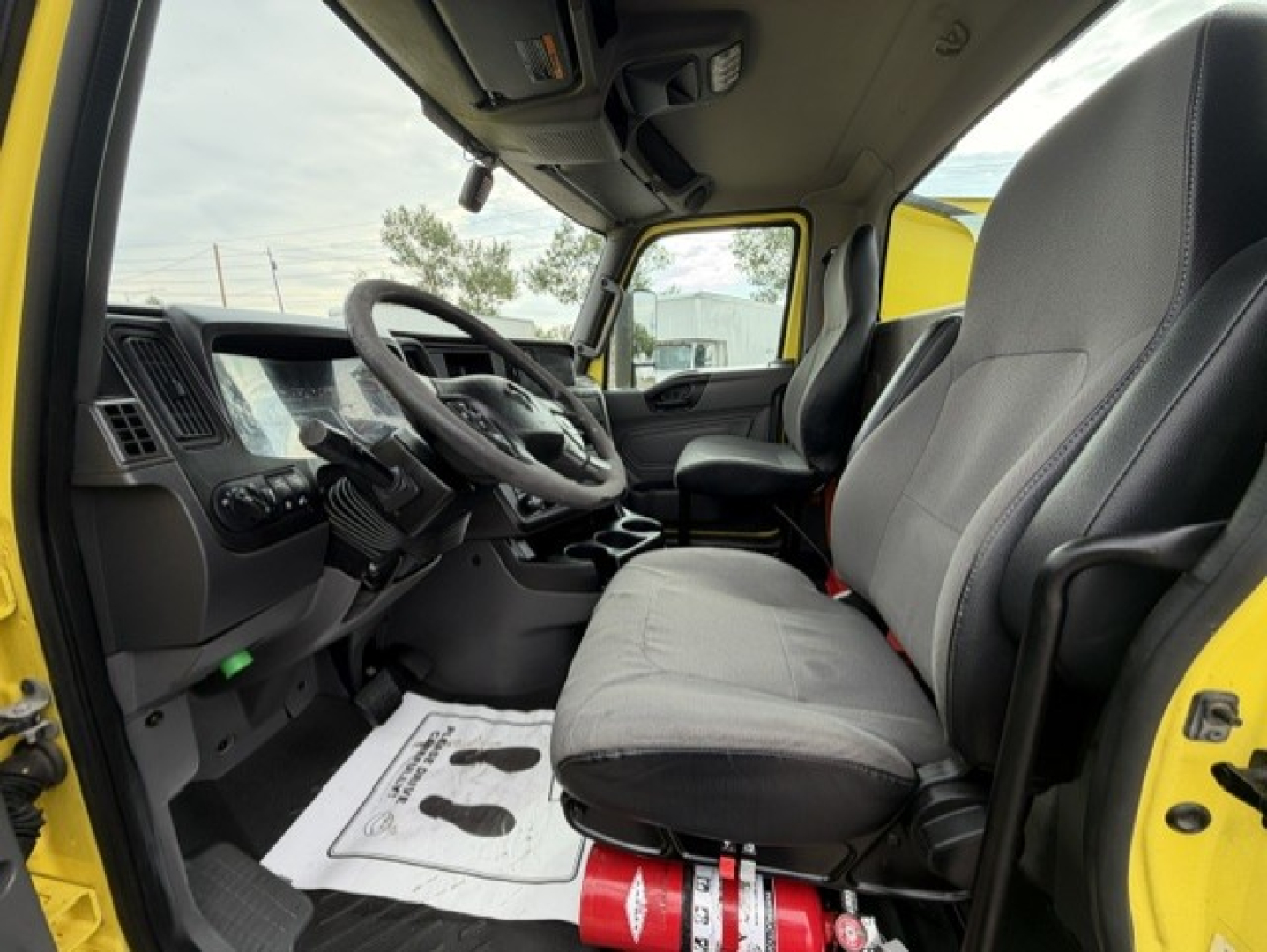 Interior view of a 2023 International LT625 daycab truck cab showing steering wheel, driver seat, and passenger seat with floor mat and a red fire extinguisher nearby.