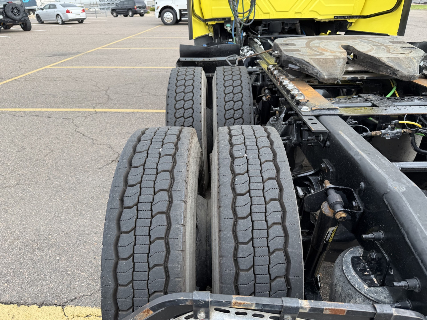 Tractor-trailer rear wheel assembly showing dual rear axles with tires and suspension components; portion of a yellow truck cab visible in background.