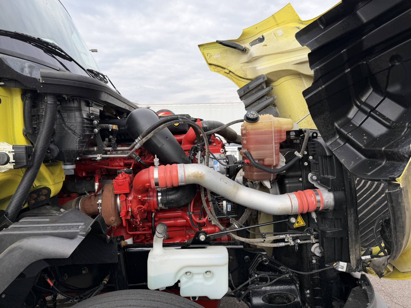 Engine bay of a 2023 International LT625 daycab showing red engine, intercooler piping, coolant reservoir, and yellow cab hood in view