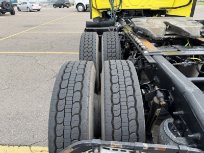 Tractor-trailer rear wheel assembly showing dual rear axles with tires and suspension components; portion of a yellow truck cab visible in background.