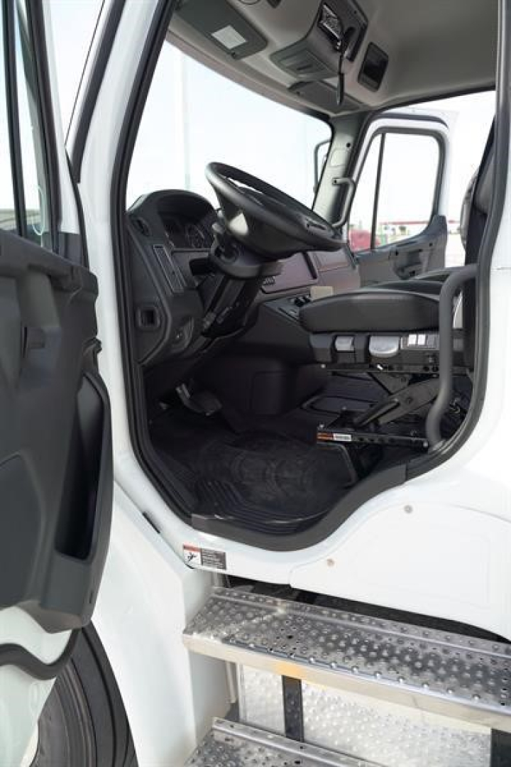 Truck cab interior view reaching from open driver's door showing steering wheel, dashboard, and entry steps of a Freightliner M2 106 Plus cab chassis.