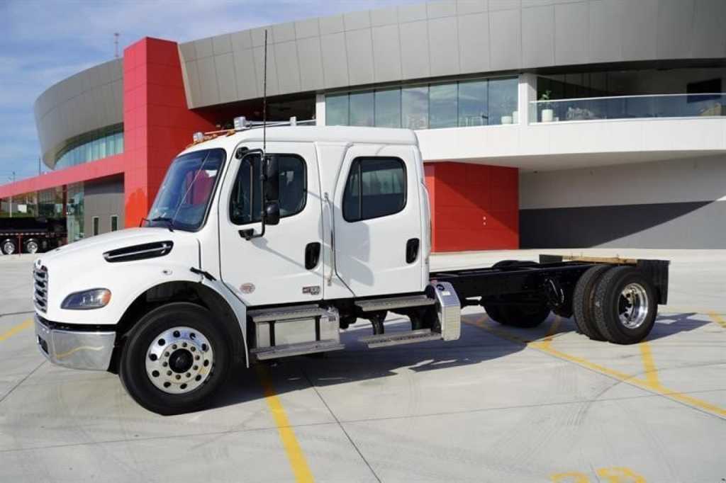 Truck cab and chassis showing Freightliner M2 106 Plus with white dual cab and chassis frame in a dealership lot.