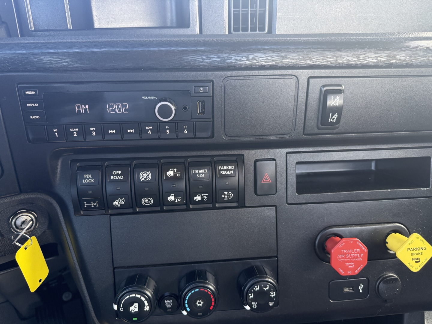 Trucks cab interior showing dashboard controls and ignition with yellow tag. AM/FM radio display and various switches and knobs. Red and yellow hazard/parking brake knobs visible.