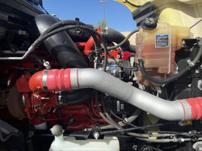 Engine bay of 2023 International LT625 showing coolant reservoir, red engine components, large intake hose, and metal piping.