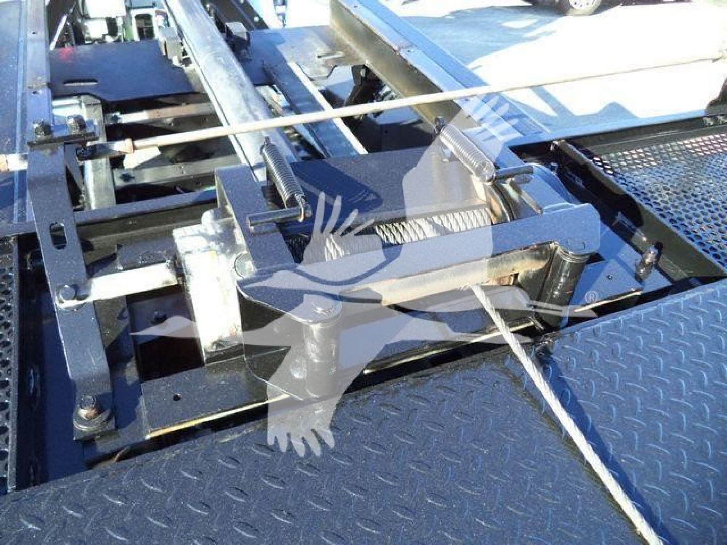 Roll-back truck chassis with linkage and hydraulic components; metal platform with diamond plate pattern and towing/coupling hardware visible.