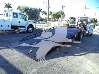 Vehicle flatbed rollback tow truck with hydraulic ramp in foreground and other trucks in background.