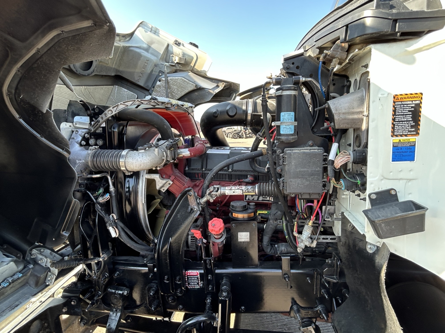 Truck engine bay view of a 2020 Mack PI64T showing engine components, hoses, reservoir caps, and battery area.