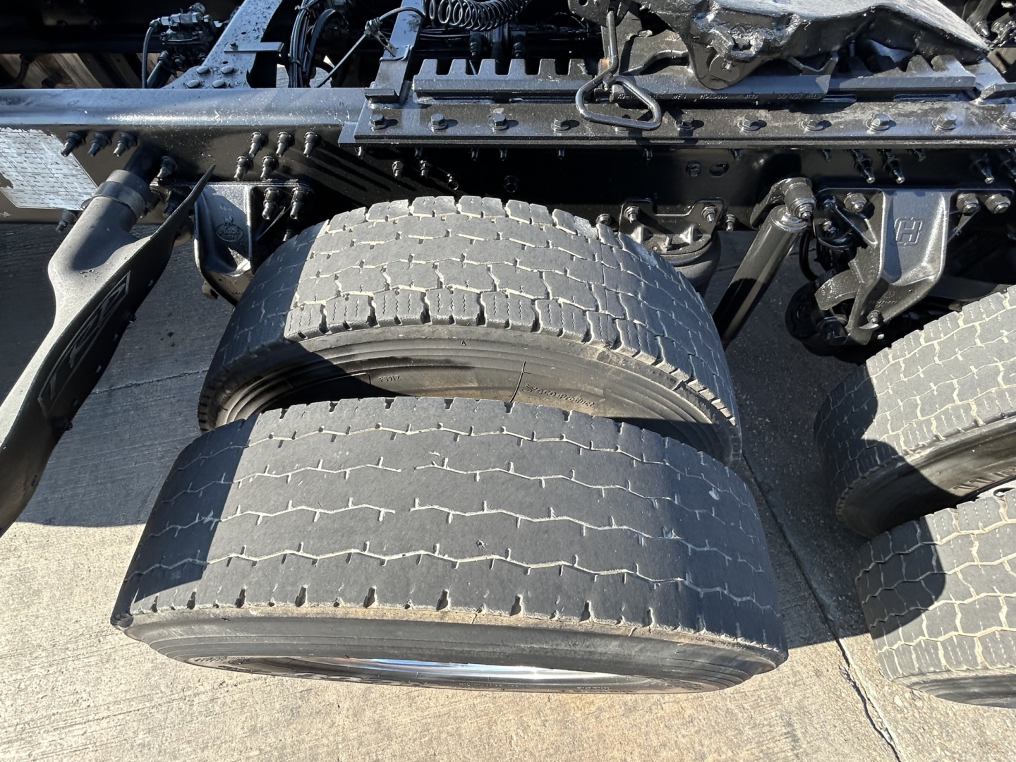 Truck wheel area showing dual rear axle tires on a Mack PI64T tractor, with visible frame, suspension components, and tire treads.